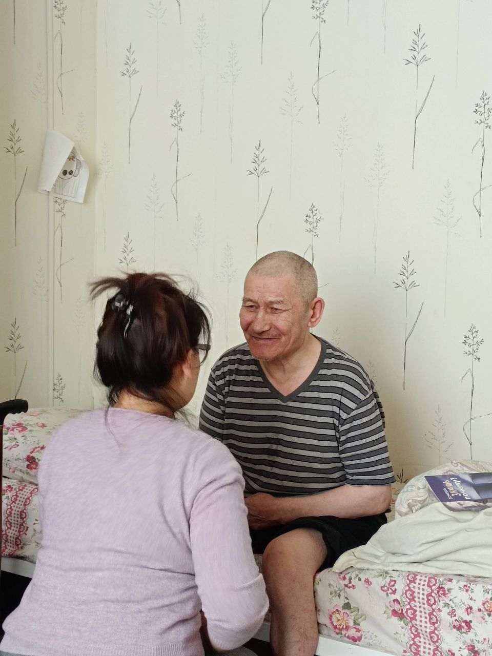 Two people sitting on a bed, talking in a small bedroom with floral wallpaper
