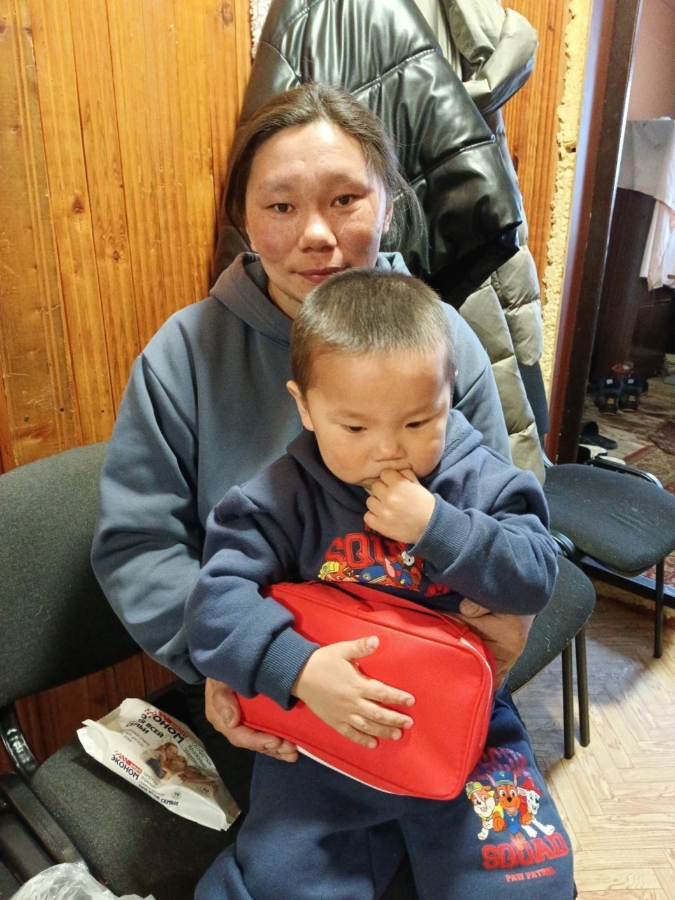 Person holding a toddler in a small indoor room, seated by a wood-paneled wall, both wearing dark clothing.