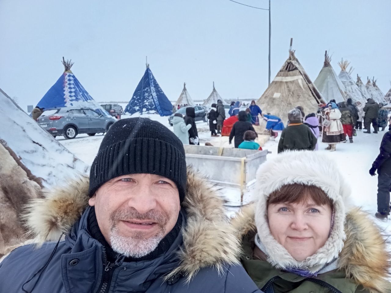 Two people in winter jackets and hats smiling at a snowy outdoor campsite with teepees and people in the background