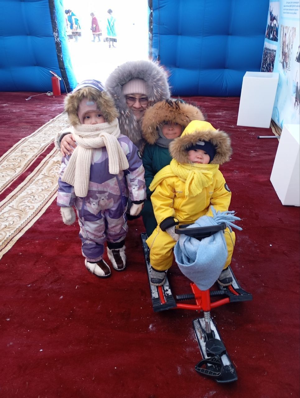 Three children in winter coats and scarves posing indoors, one seated on a red toy scooter.
