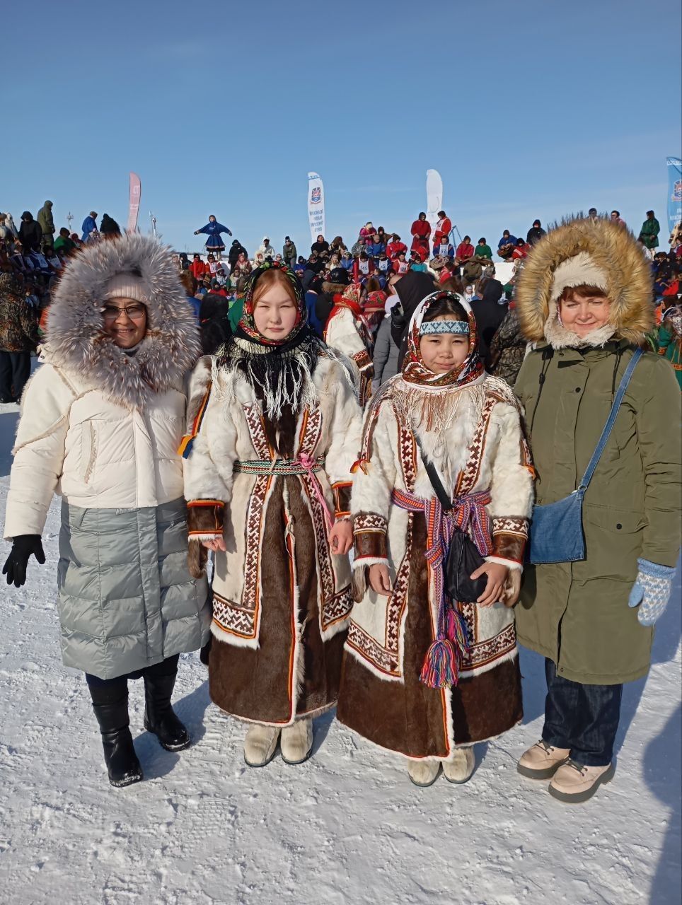 Four people in fur-lined winter coats standing together in a snowy outdoor festival crowd