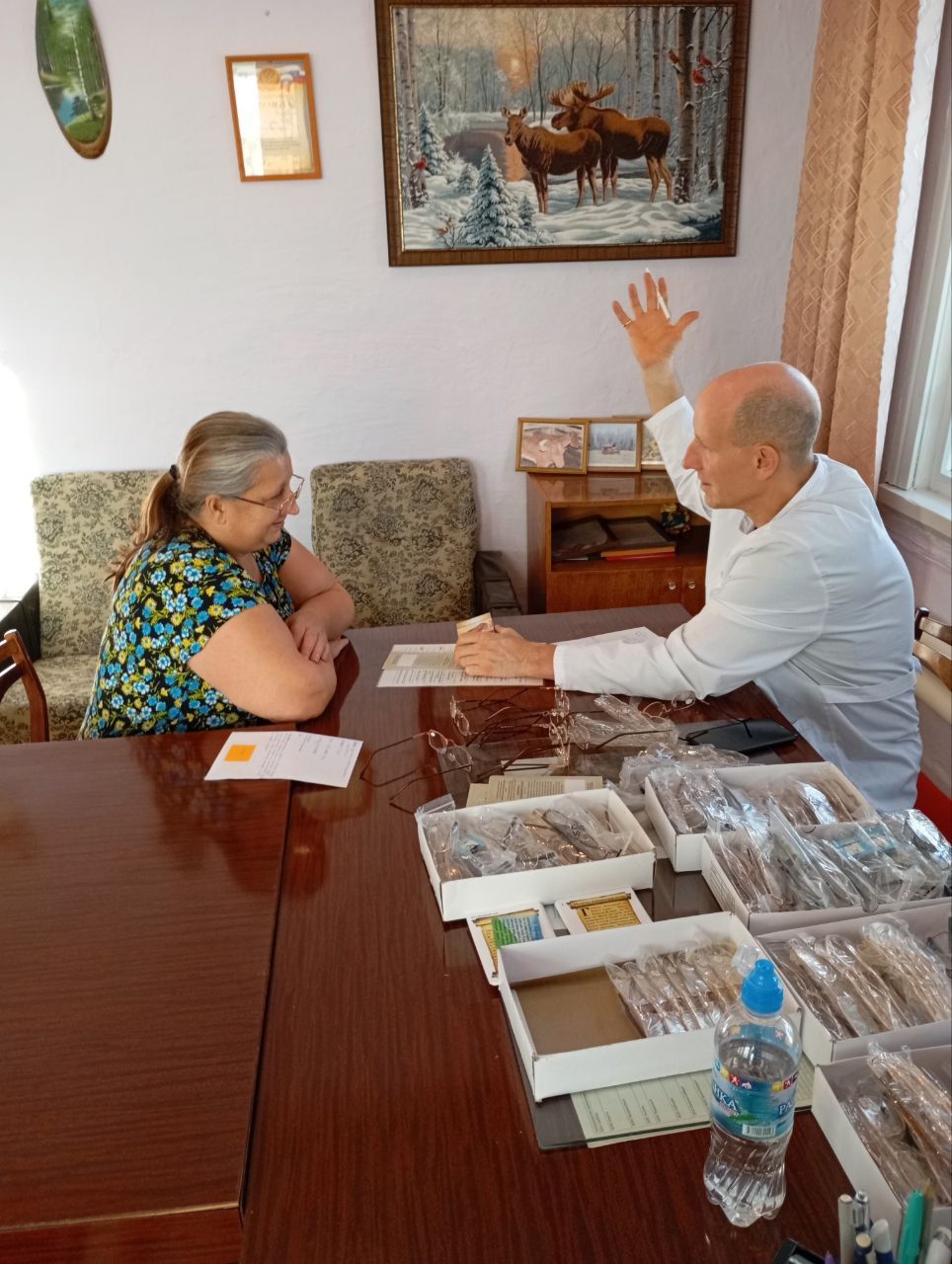 Two people seated at a table in a meeting room, talking with brochures and a water bottle on the table
