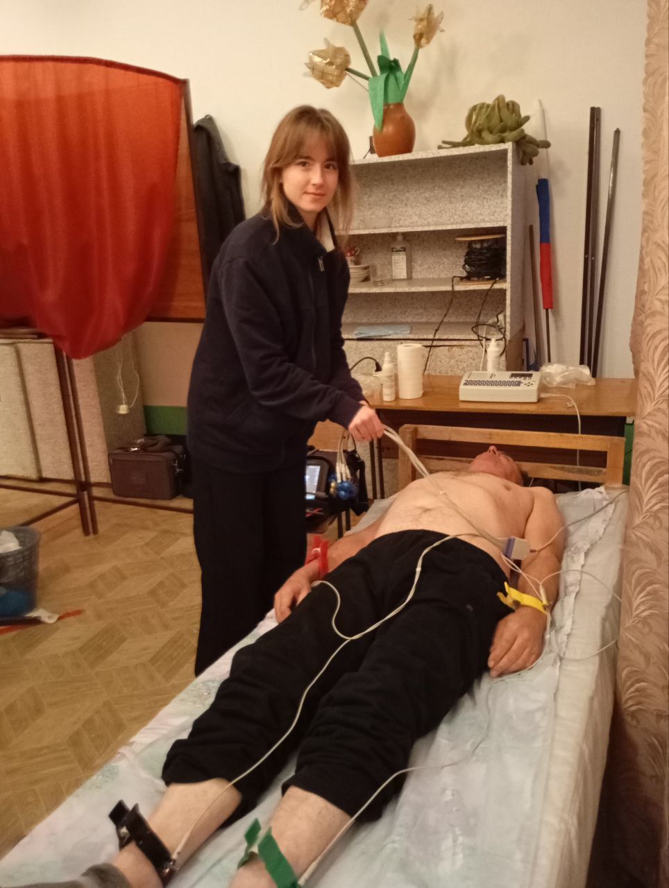 Woman standing beside a person lying on a table in a room, with wires attached and red curtain in background
