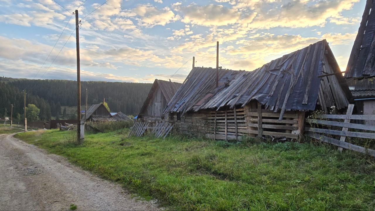 Rural dirt road beside weathered wooden huts with steep roofs under a cloudy sunset sky
