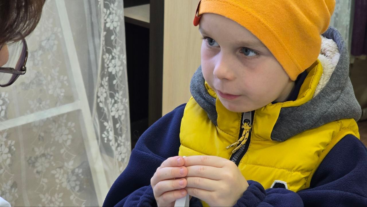 Child in orange hat and yellow vest looking at something while holding a small object indoors