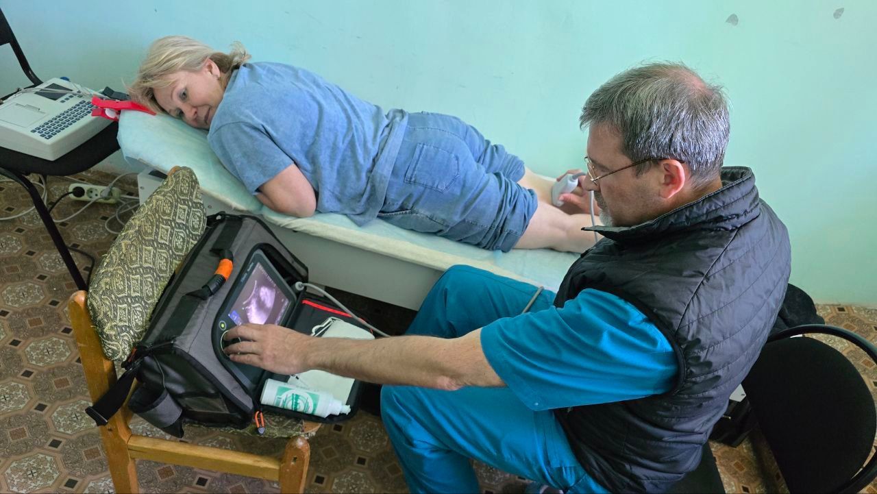 Two people in a room, one lying on a cot while another adjusts a medical device nearby.