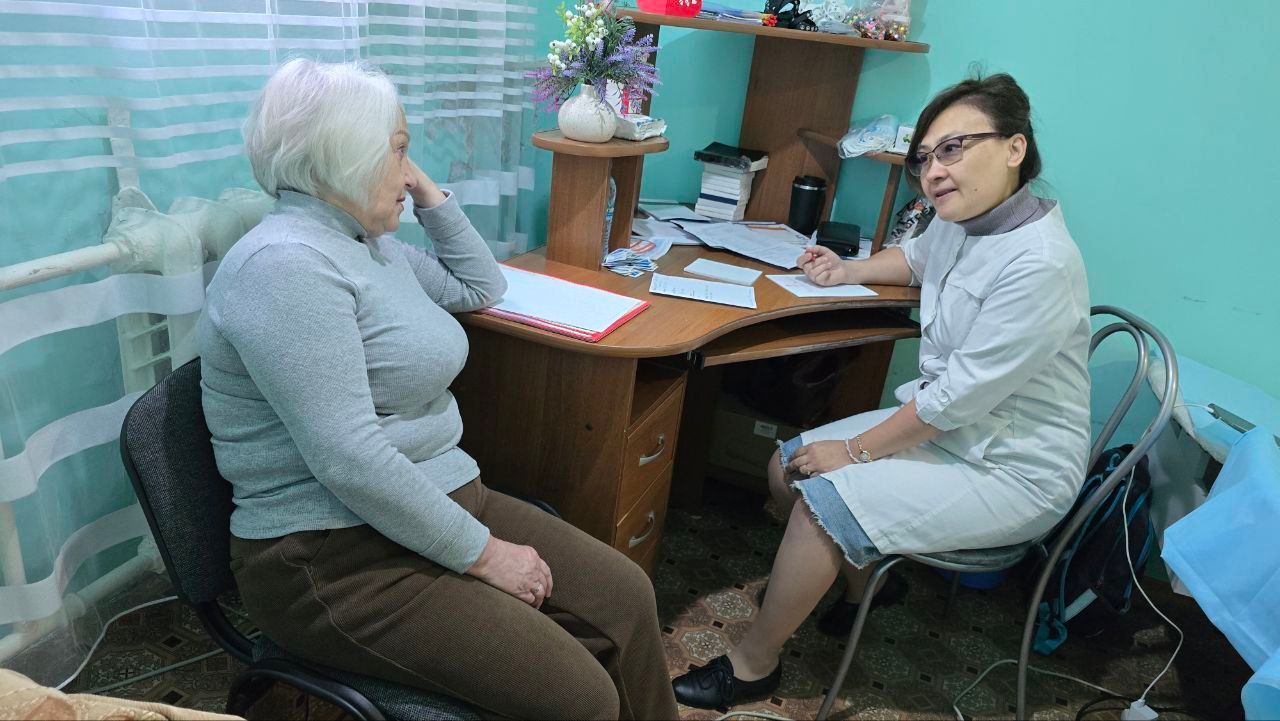 Two women sit across a desk in a small turquoise office, one writing while the other turns toward the camera.