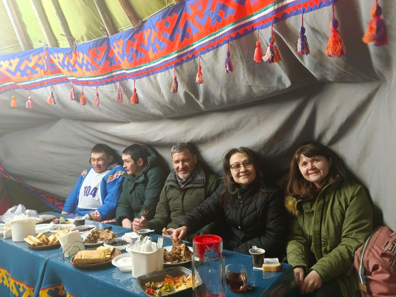 Five people seated at a table with tea and food inside a colorful tent, smiling at the camera