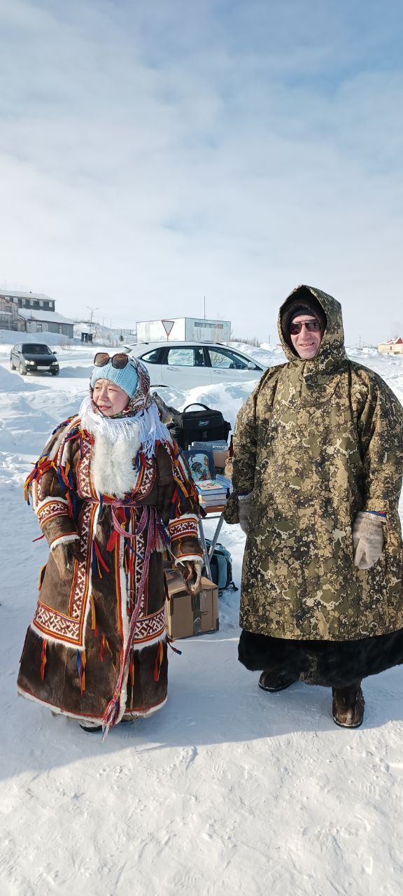 Two people in fur-lined winter coats standing in a snowy landscape with cars behind them