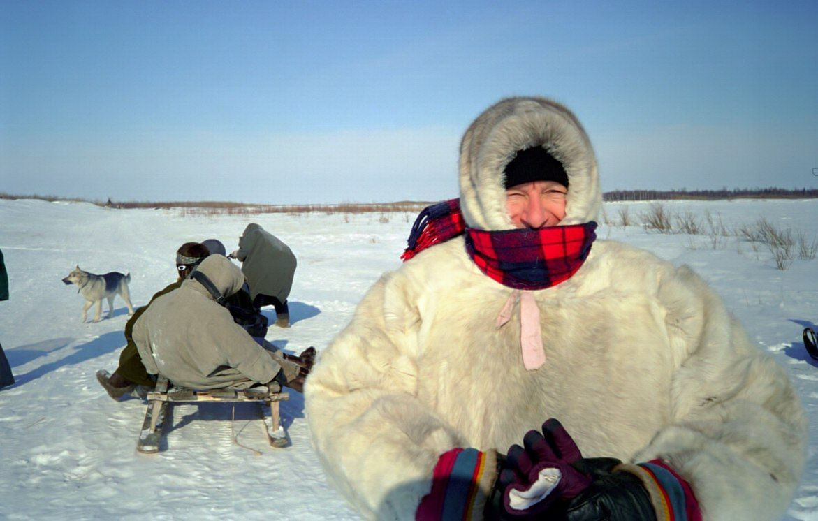 Person in a fur-lined hood and red scarf standing in a snowy field, with people sledding behind them.