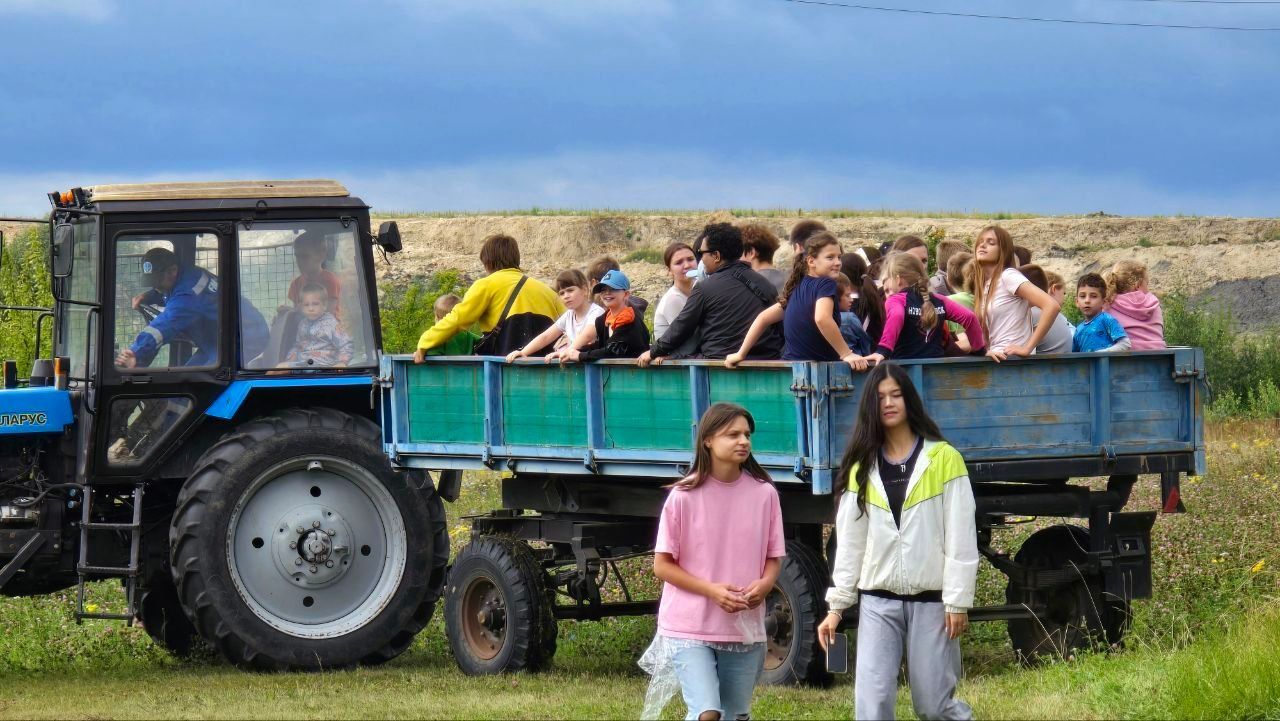 Blue tractor pulling a crowded trailer of people through a grassy field, with two women standing in front