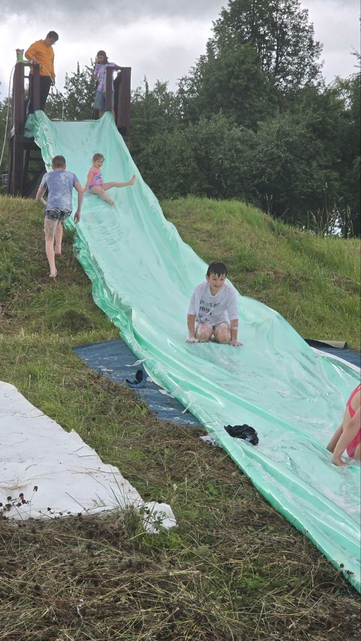 People sliding down a long green tarp slide on a grassy hill outdoors