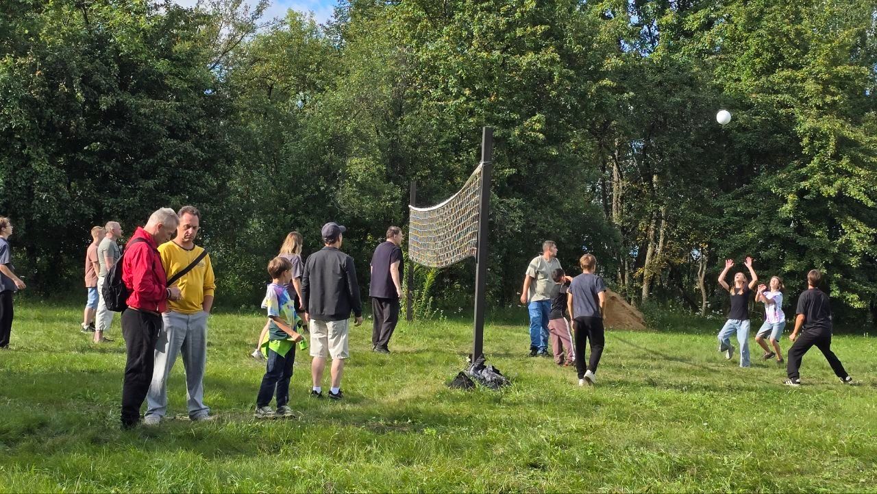 People standing and walking on a grassy field near a volleyball net in a park, with trees in the background.