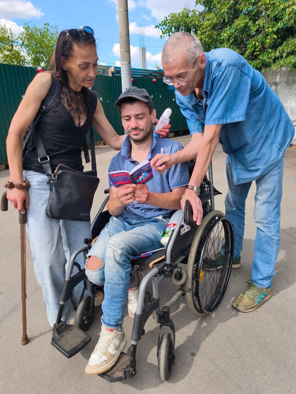 Three people gathered around a seated man in a wheelchair, looking at a phone outdoors.