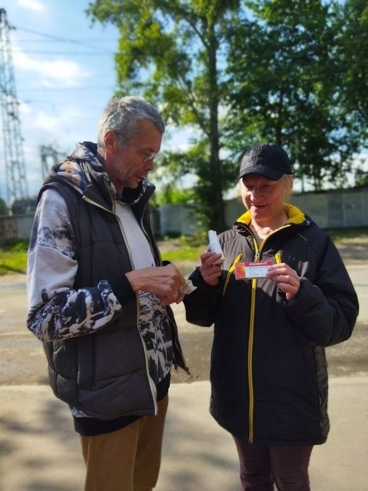 Two people outdoors examining a small item together on a sunny path, with trees and a fence behind them.