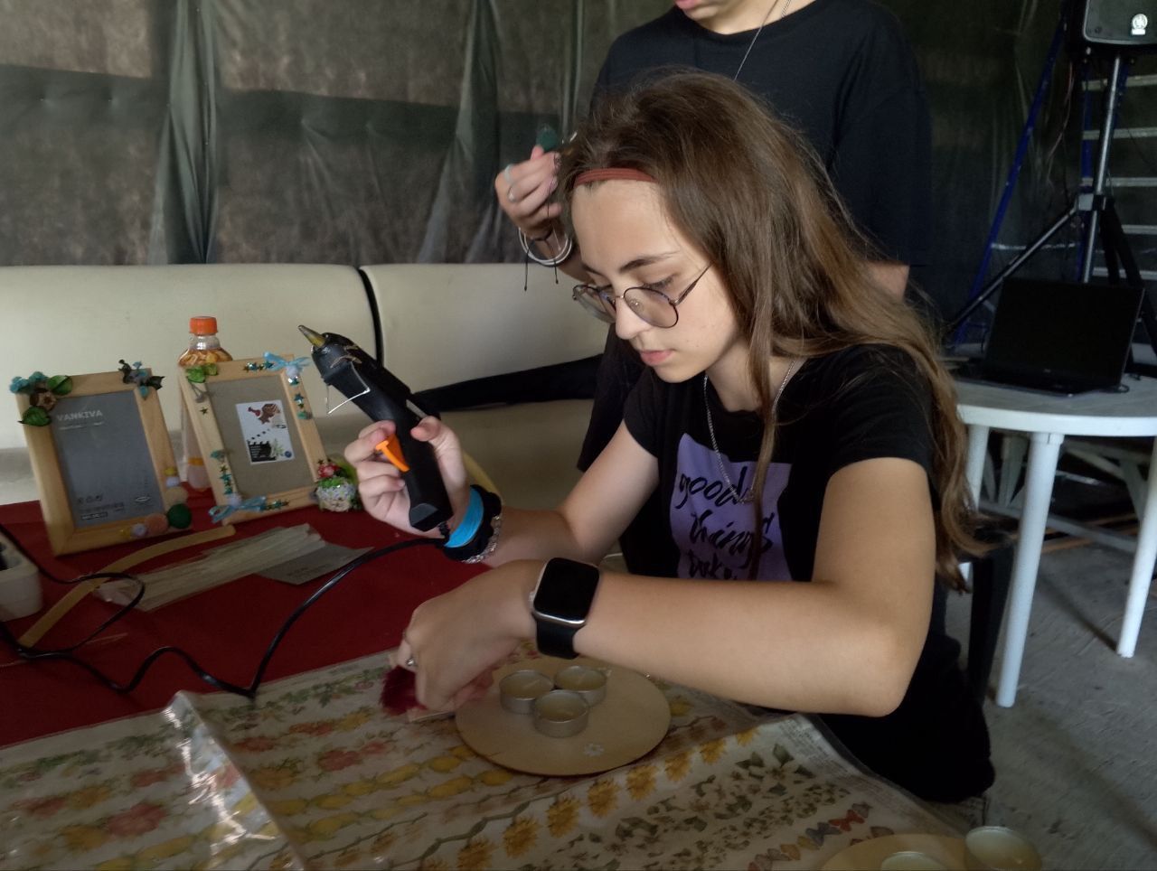 Person working at a craft table, using a hot glue gun on a circular project indoors.