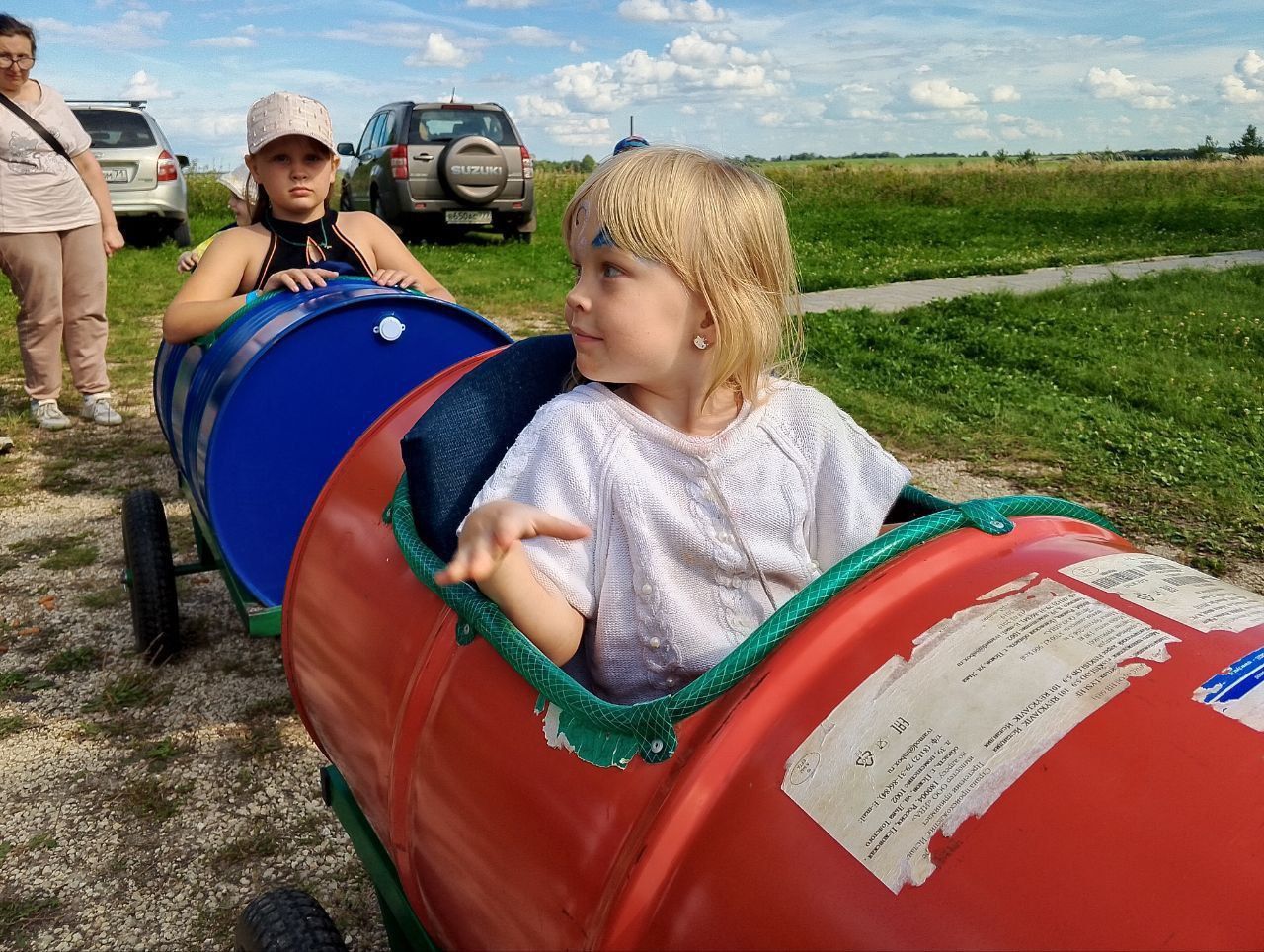 Children riding in colorful barrel train cars on a grassy field under a cloudy sky