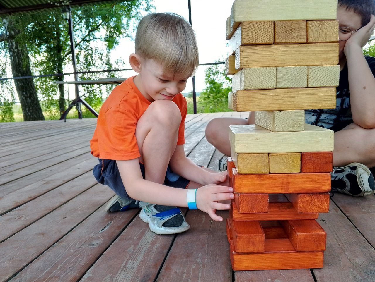 Child crouches beside a stacked block tower on a wooden deck outdoors