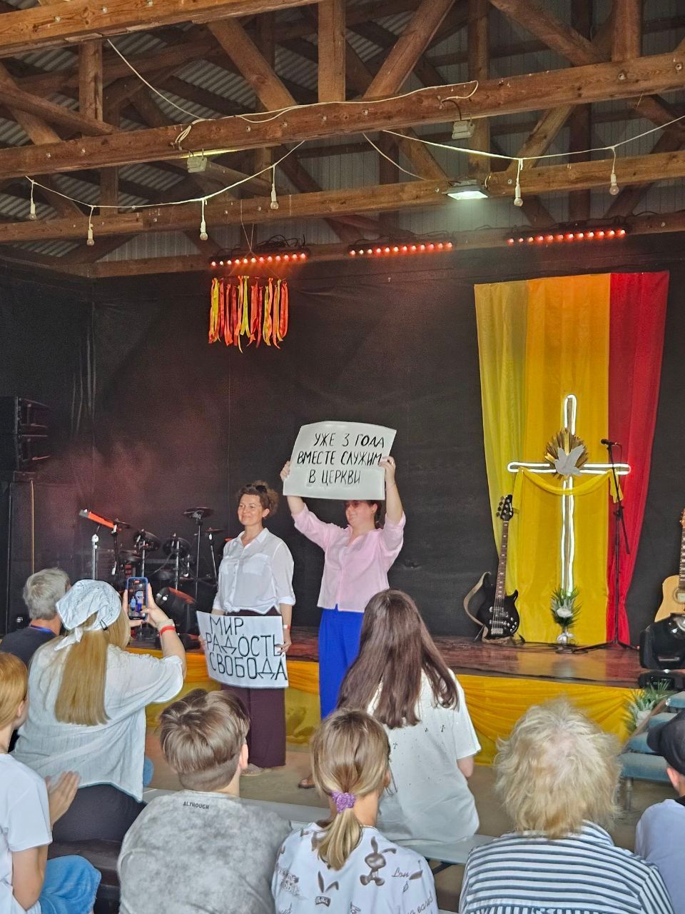 Stage performance with people holding signs and a microphone in a rustic hall, audience watching