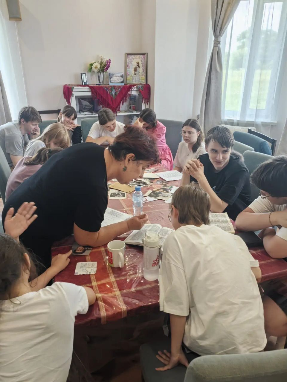People gathered around a table, sharing food in a bright room with a red cloth on a side table.