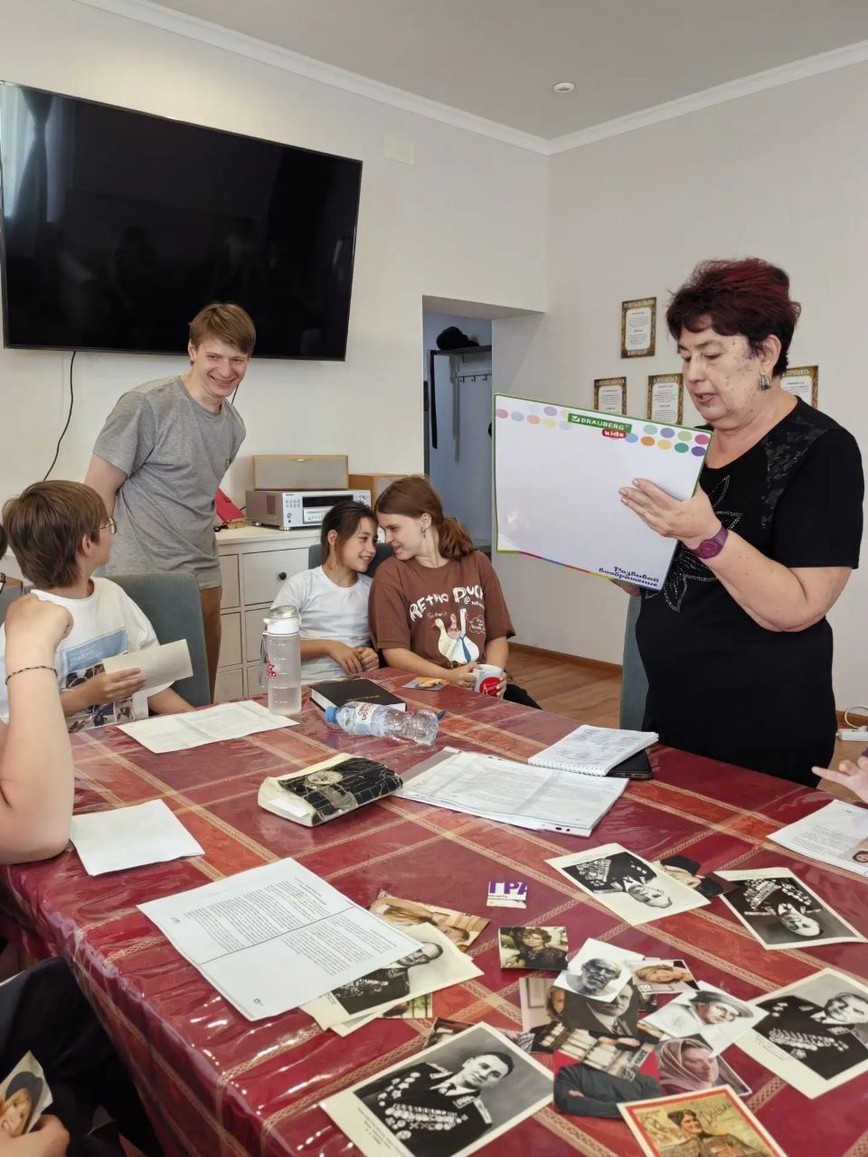 People gathered around a table with papers and photos, while a woman presents from a clipboard in a meeting room
