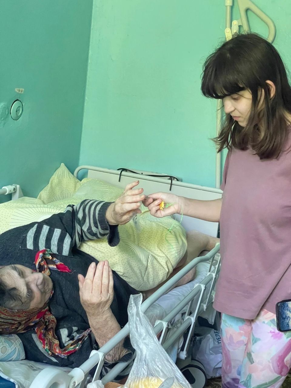 Woman standing beside a bed reaching toward an elderly person lying in a hospital-style bed
