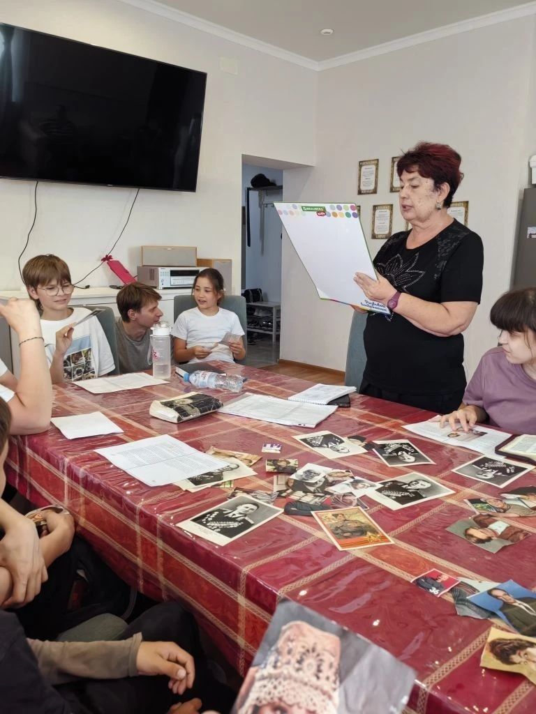 Teacher reading from papers to a group around a table in a classroom with photos and documents.