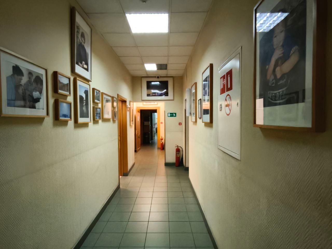 Narrow hallway with framed posters on beige walls and tiled floor, lit by fluorescent ceiling lights.