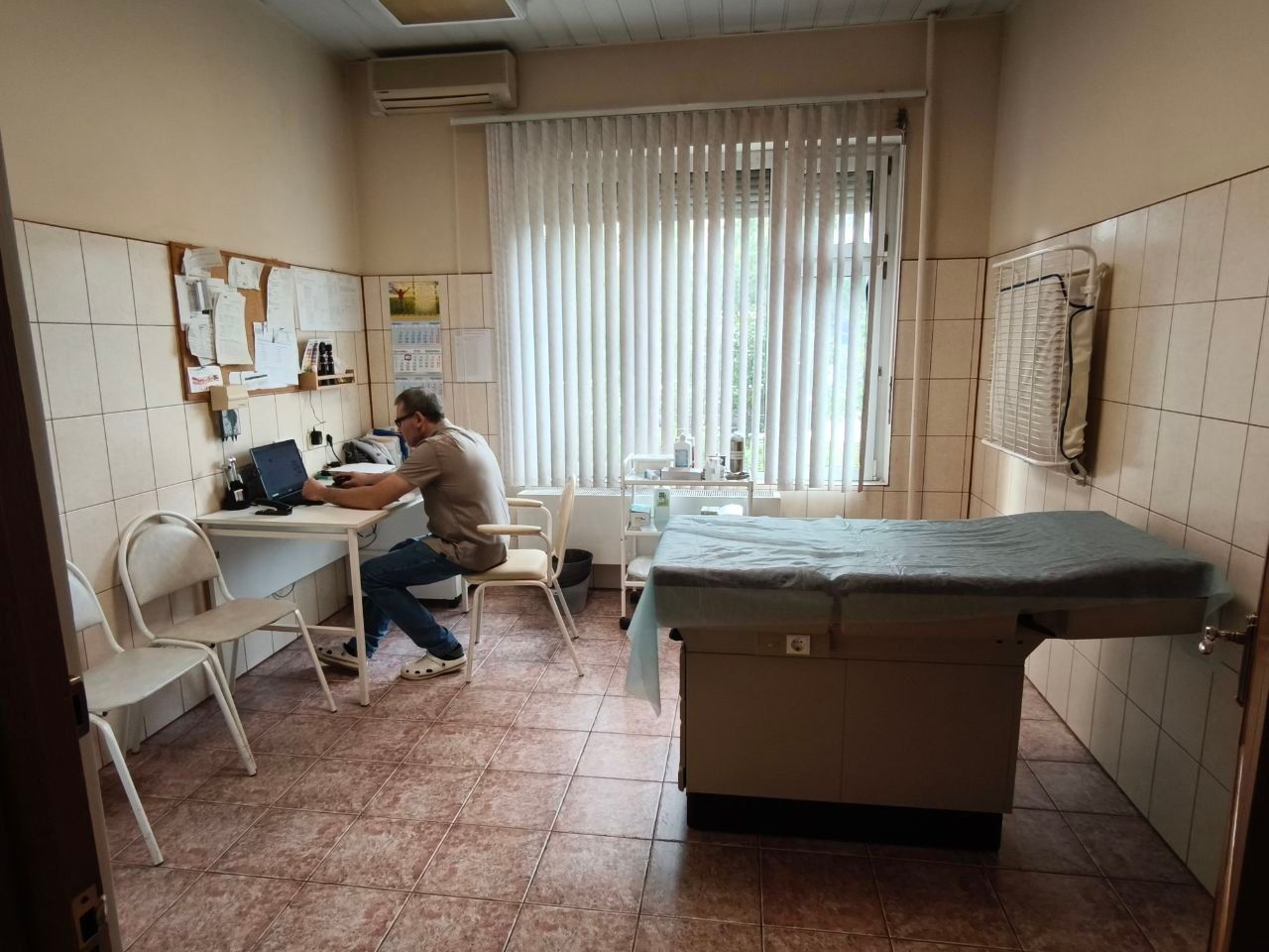 Medical exam room with a person seated at a desk and an exam table under a window