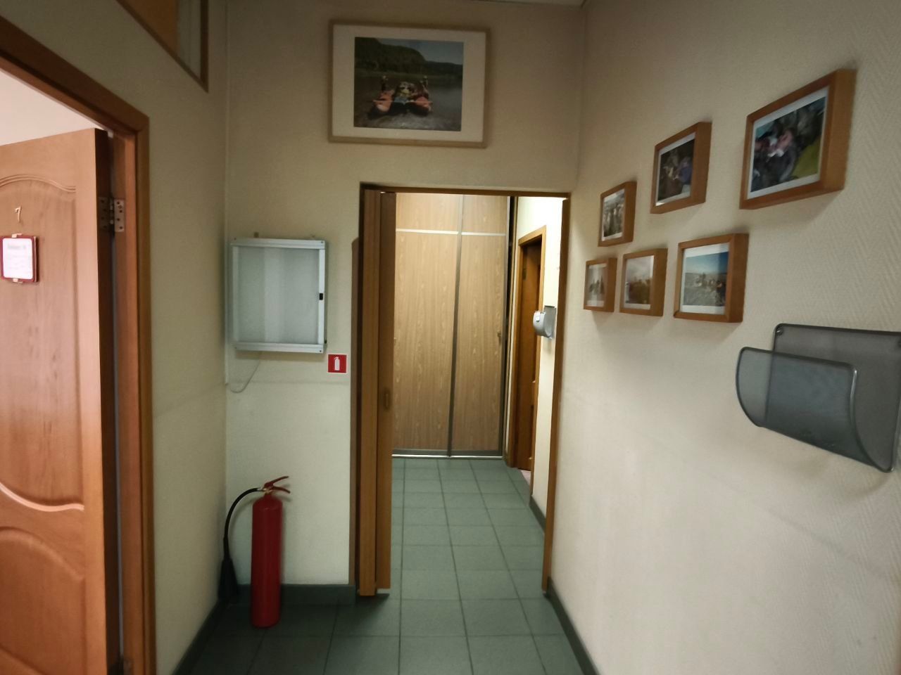 Narrow hallway with beige walls, tiled floor, wooden doors, framed pictures, fire extinguisher, and wall cabinet.