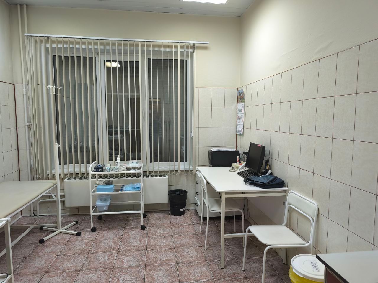 Small clinical room with bed, desks, chair, computer, and barred window blinds