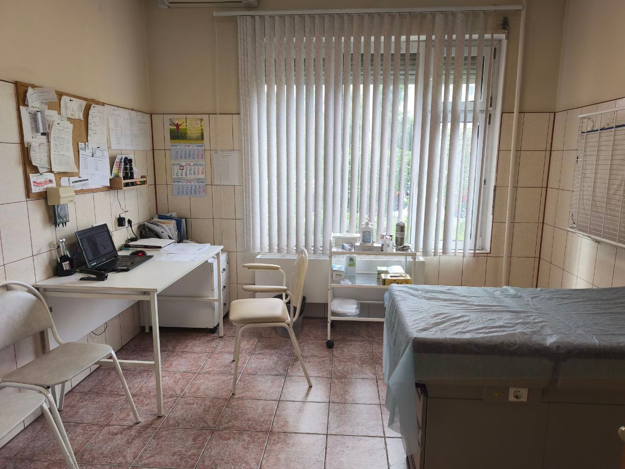 Small clinic exam room with desk, chairs, bulletin boards, and an exam table by a window with blinds