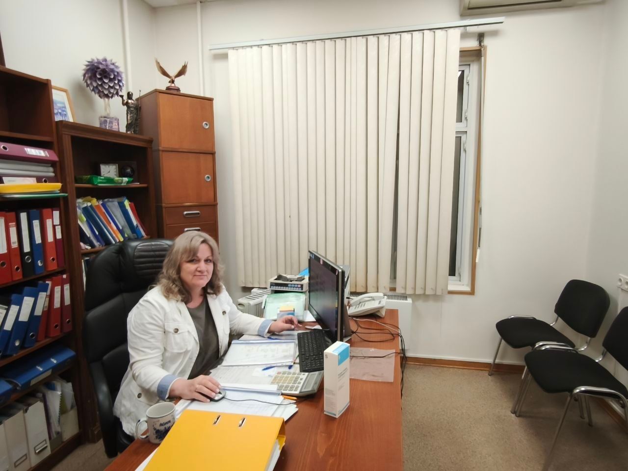 Woman seated at an office desk with computer and papers, in a small room with shelves and chairs