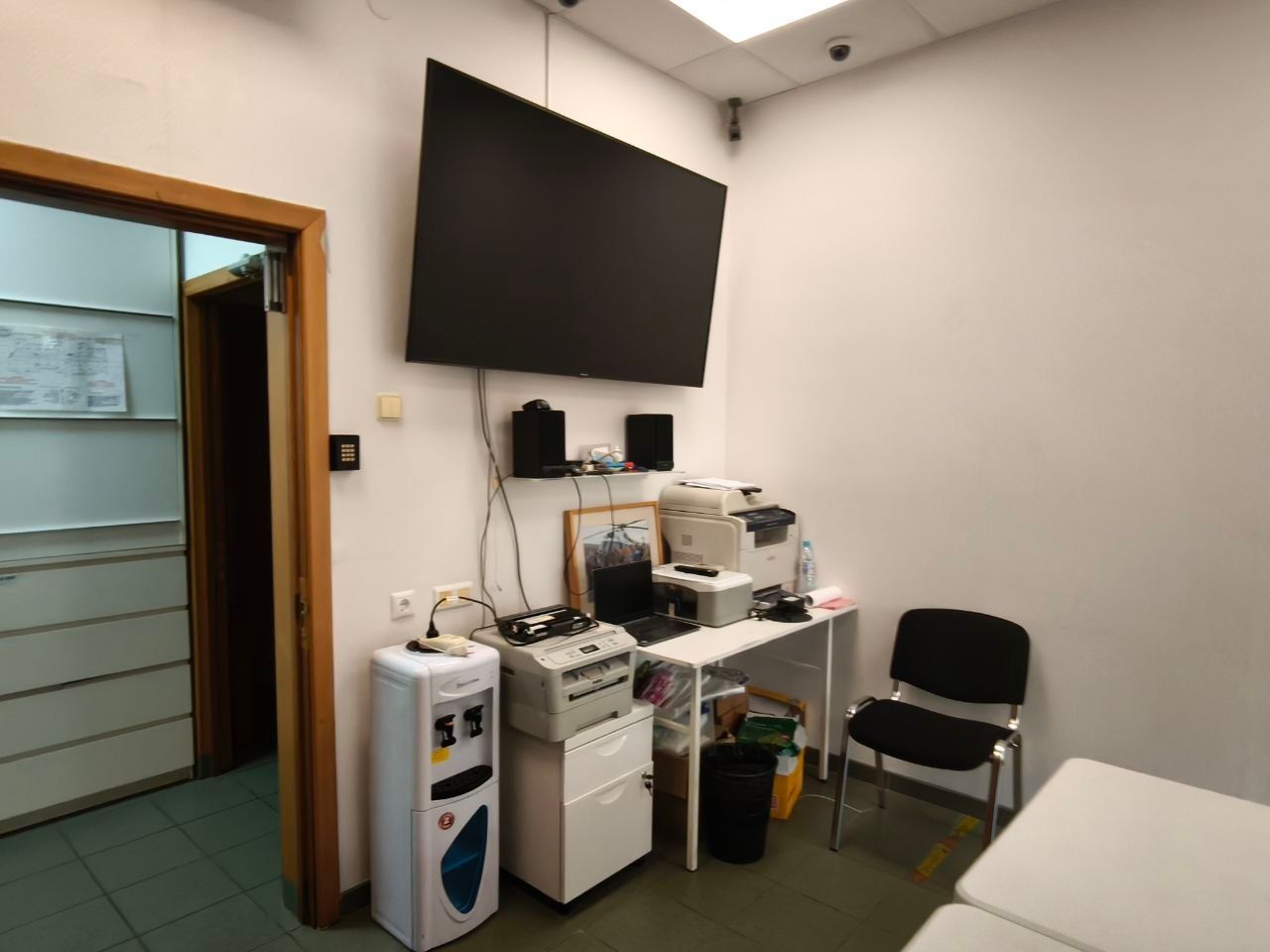 Small medical exam room with wall-mounted TV, equipment cart, chair, and bed.