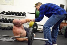 A man is helping another man lift a barbell in a gym.