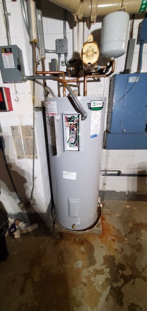 A water heater in a basement. The floor has rust-colored stains. Piping is above the tank. Electrical components are visible.
