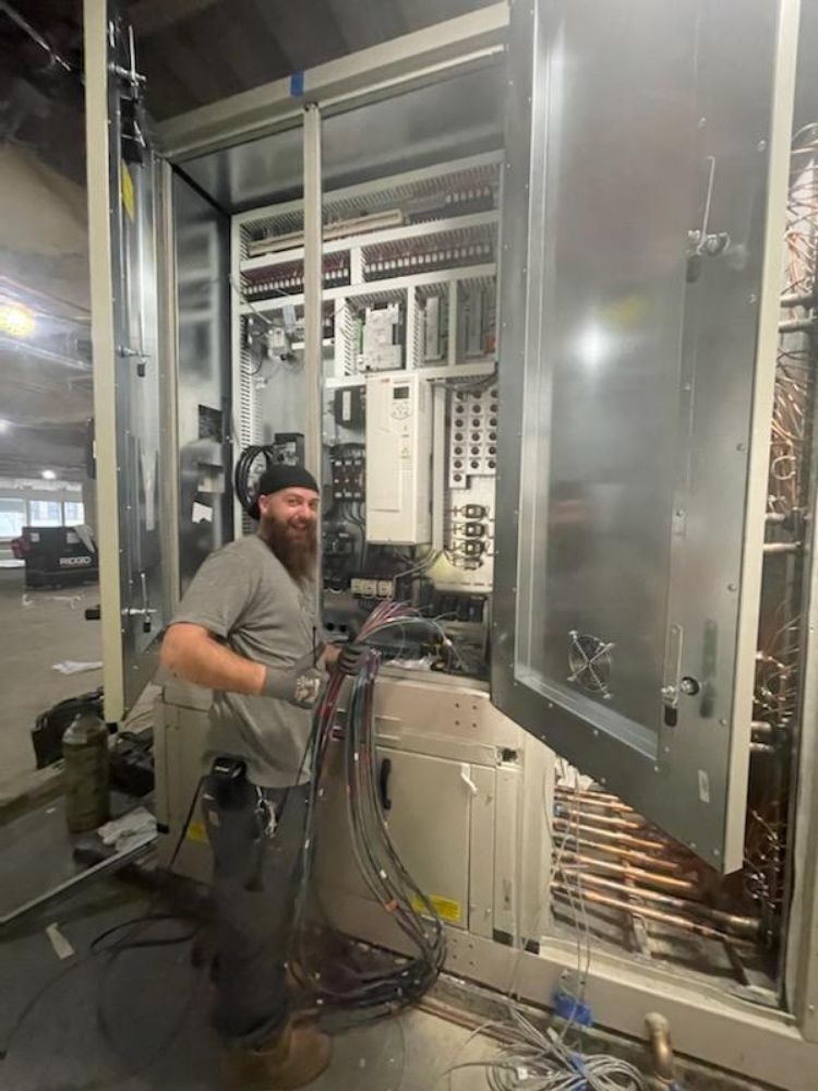 Man in a gray shirt working on large electrical equipment. He has a beard and is smiling. Interior setting.