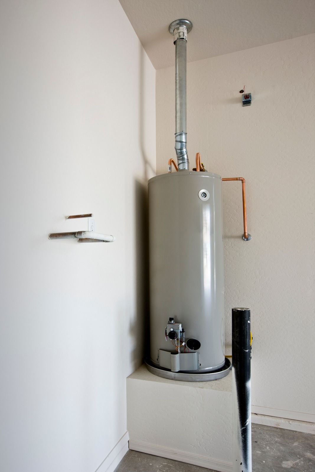 Water heater in a corner, with vent pipe. White walls and small concrete base.