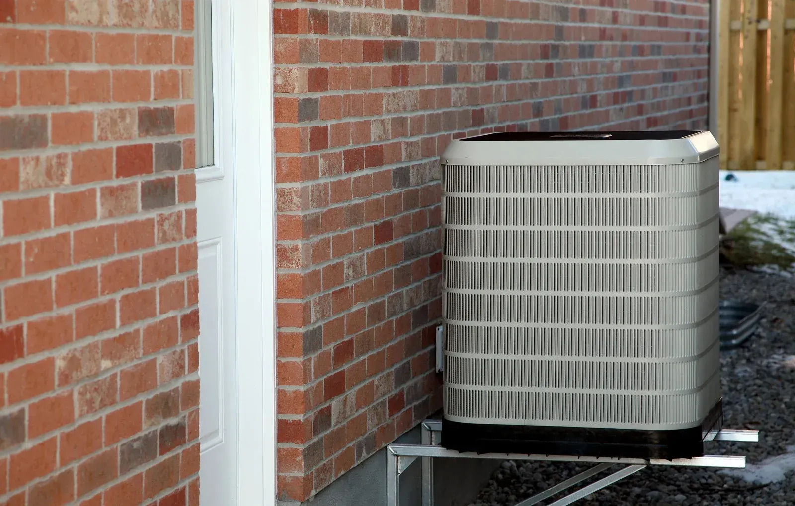 Air conditioner unit beside a brick wall, resting on a metal stand outdoors.