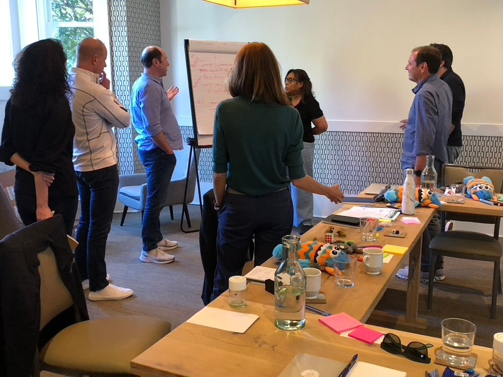 People standing and talking in a meeting room beside a table with drinks, snacks, and notes.