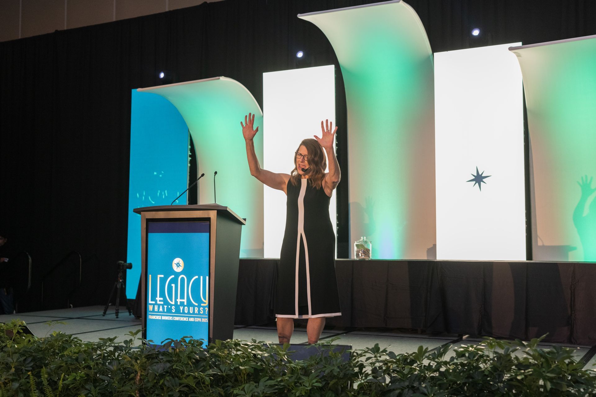 Speaker on stage beside a podium, arms raised before a teal-and-white conference backdrop.
