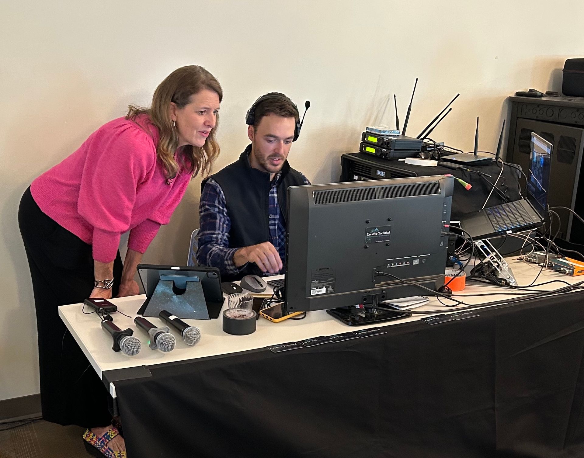 Two people lean over a desktop computer on a cluttered table with cables and equipment.