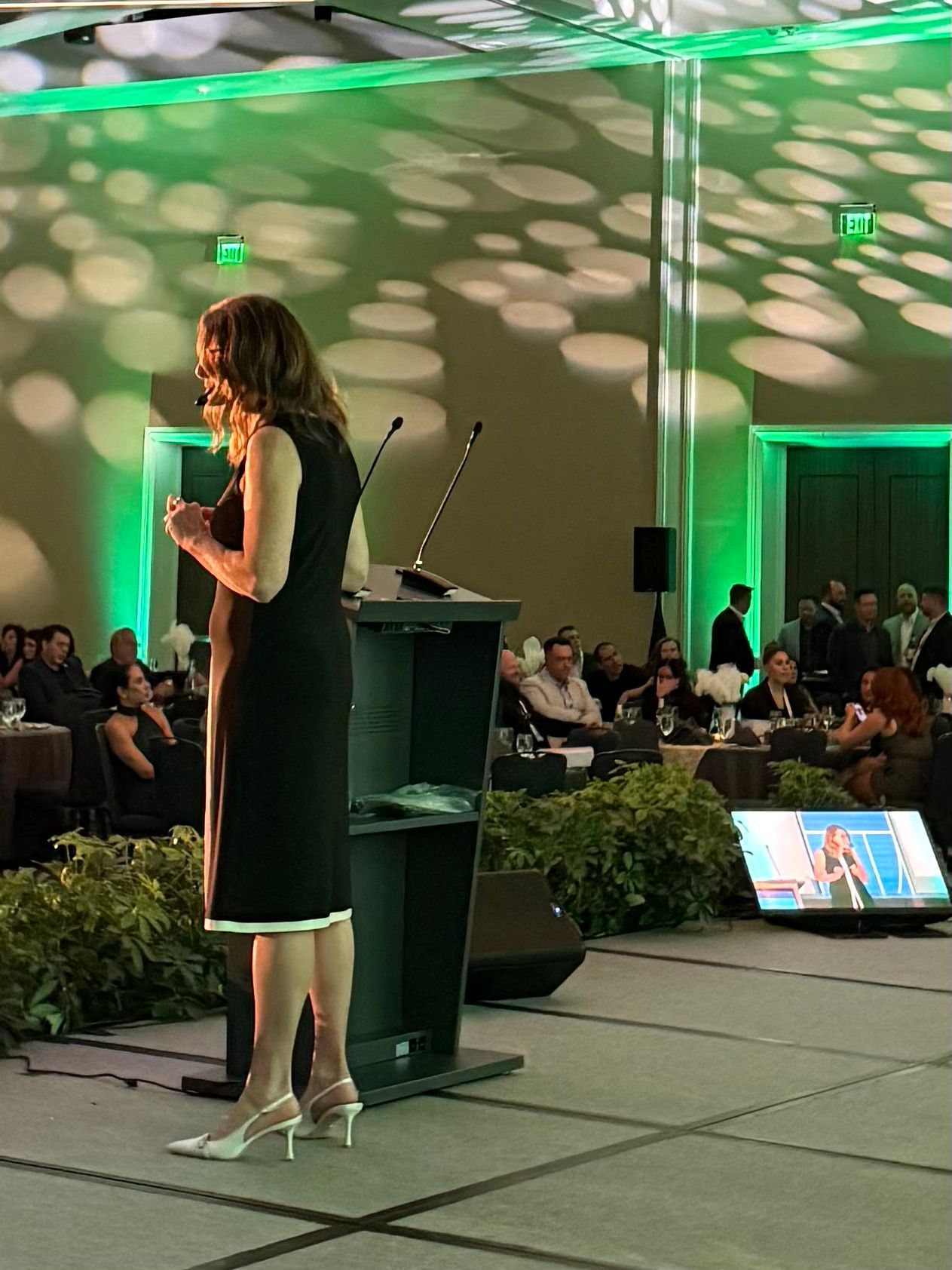 Speaker in black dress presents at a podium in a banquet hall with green lighting and seated audience.