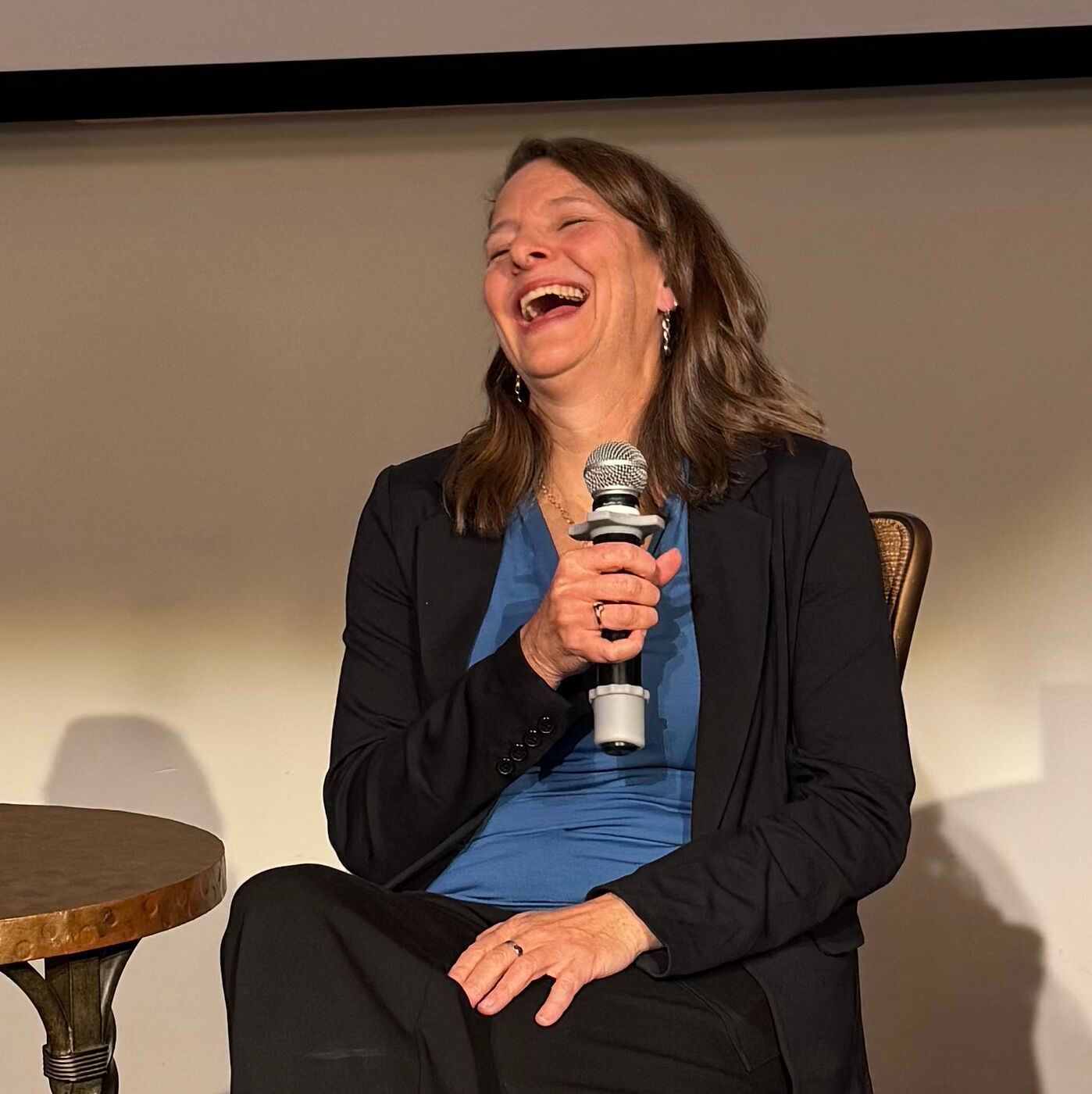 Person laughing while holding a microphone on a stage, seated in a chair beside a small table.
