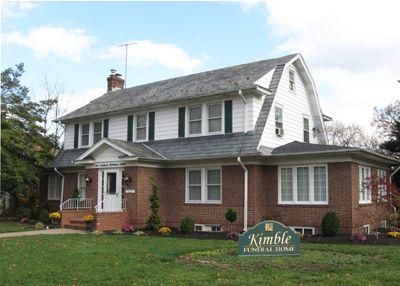 Two-story brick house with white siding and black shutters, set on a lawn with a sign in front.