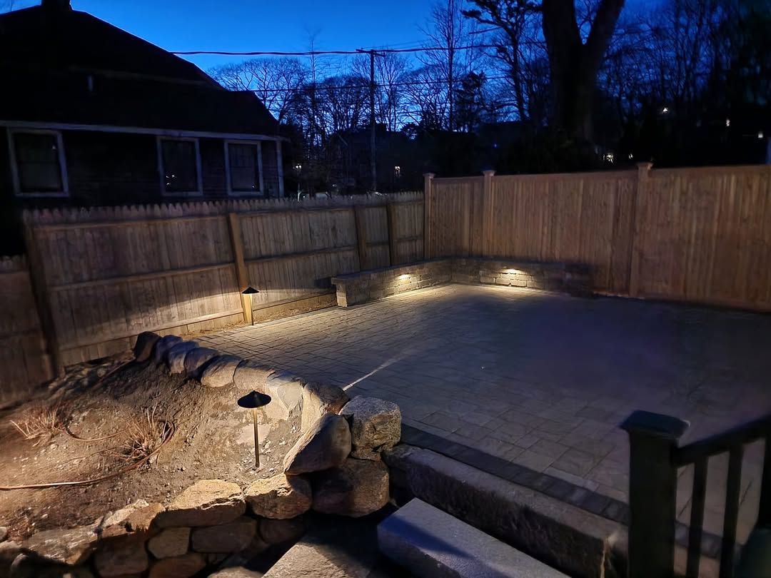 Outdoor patio with cedar fence, stone wall, and in-ground lighting at dusk. Custom nightlighting