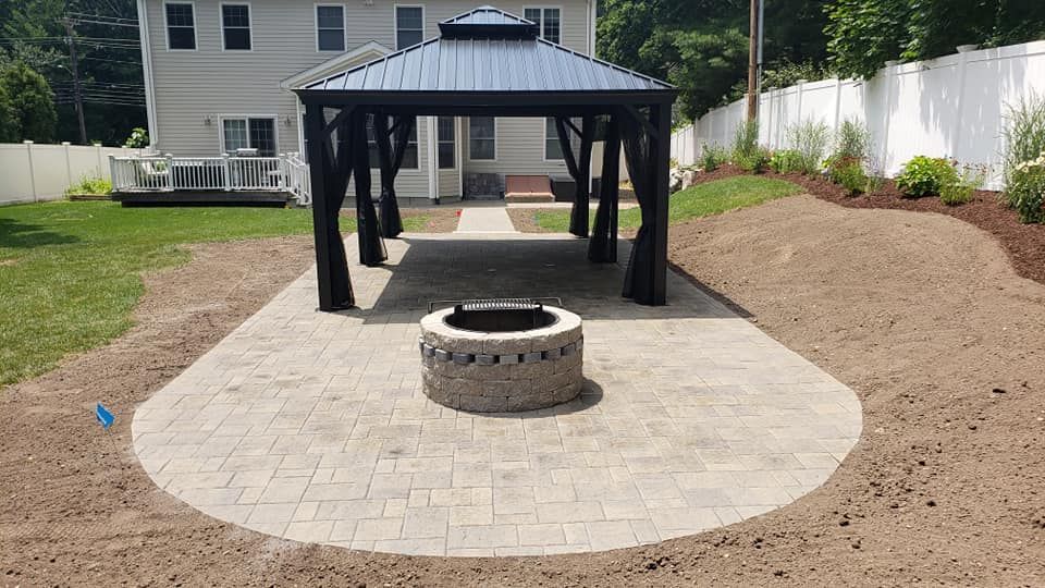Patio with a gazebo, fire pit, and home in the background. Brown brick pavers and freshly laid mulch.