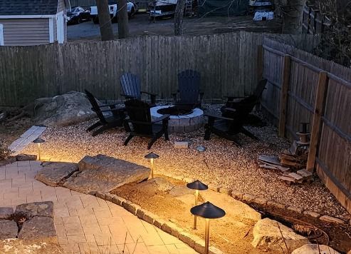 Backyard fire pit area with Adirondack chairs, gravel, and pathway lighting.