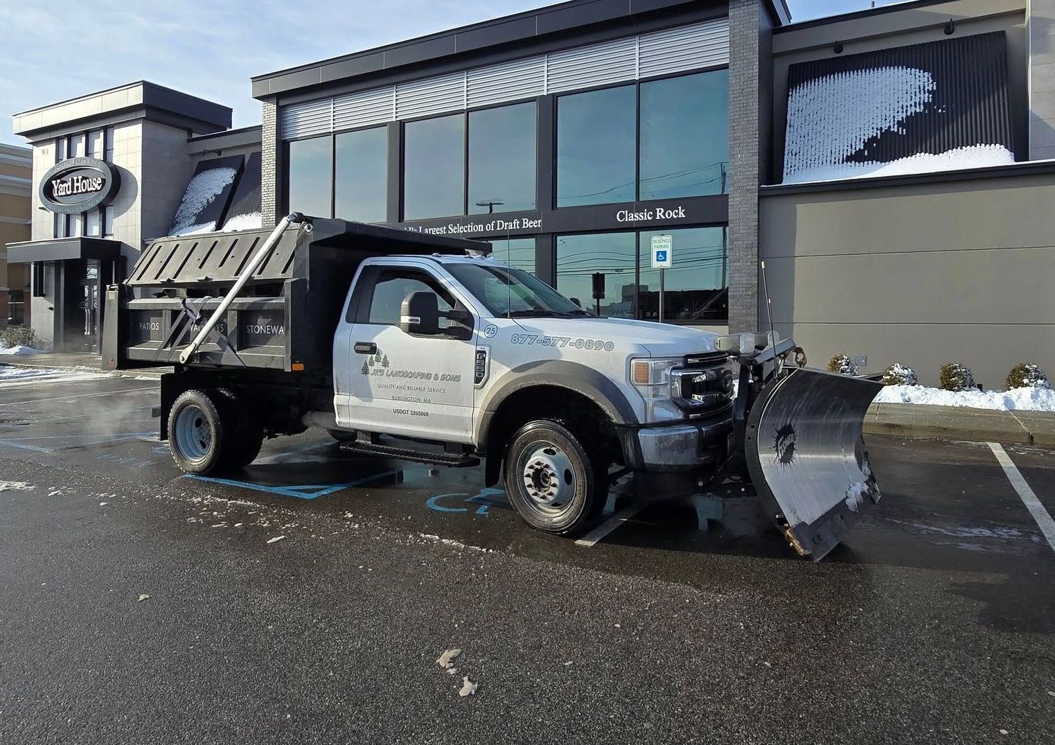 Snowplow truck clearing a parking lot in front of a modern building with large windows on a sunny day.