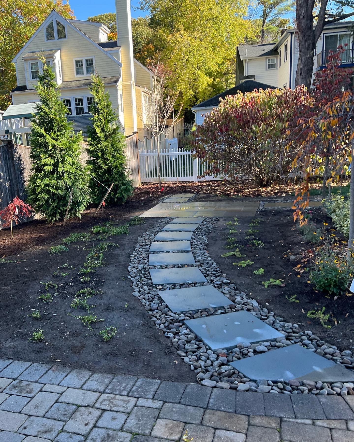 Stepping stone path curves through a garden with a white picket fence, flanked by trees and a bluestone patio.